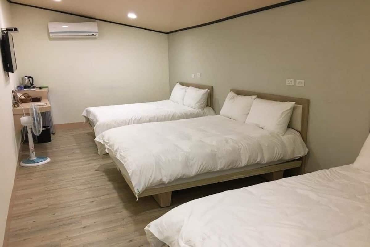 A hotel room with two single beds, a desk with a chair, a standing fan, and an air conditioning unit.