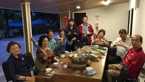 Un groupe de personnes est assis autour d’une table en bois, devant divers plats, dans une pièce dont le plafond est également en bois et qui offre une vue sur les arbres à l’extérieur.