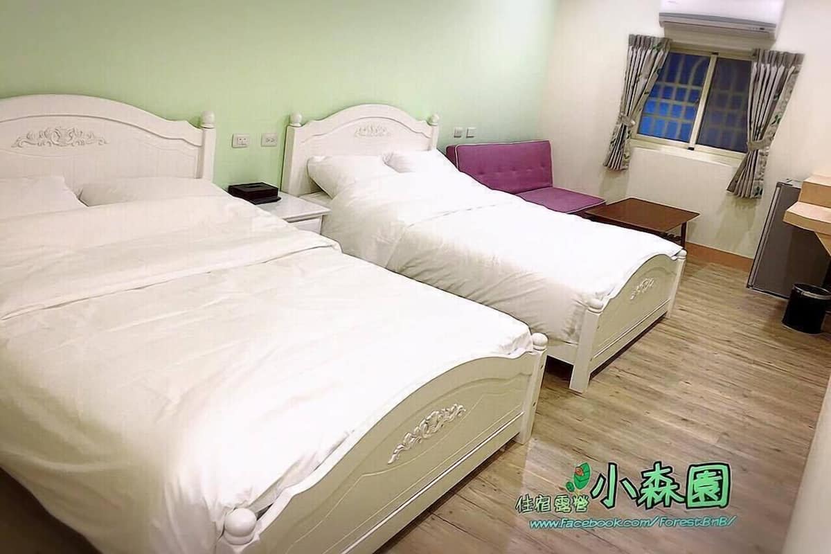 A hotel room with two beds, a small table, a chair, an air conditioning unit, and a window with curtains.