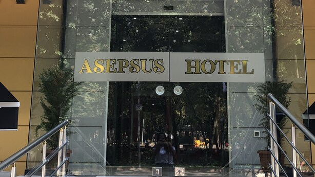 La entrada del Hotel Asepsis, con fachada de vidrio, un conjunto de escaleras que conducen a la entrada y una fila de banderas en la parte superior.