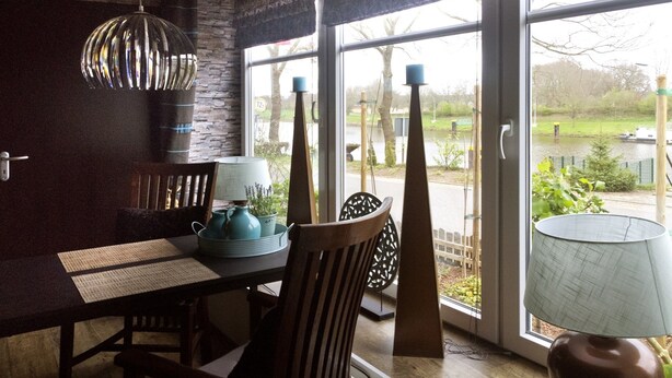 A dining area with a table, chairs, a lamp, and a view of a river and greenery outside. - Gasthaus Düther Schleuse (Fresenburg)