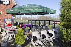 An outdoor seating area with a green umbrella, white plastic chairs, and potted plants. - Gasthaus Düther Schleuse (Fresenburg)