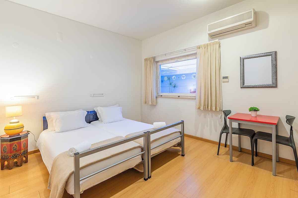 Ein Hotelzimmer mit Bett, Nachttisch, Lampe, Fenster, Klimaanlage und einem kleinen Tisch mit Stuhl.