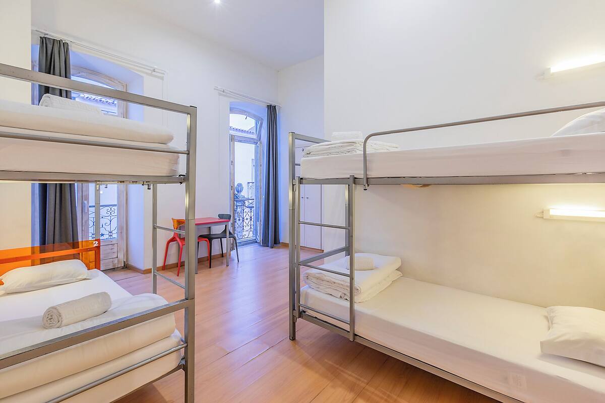 Ein Schlafsaalzimmer mit Etagenbetten, einem kleinen Tisch, Stühlen und einem Balkon mit Geländer.