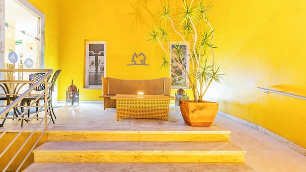 A bright yellow interior with a wicker sofa, a dining table with chairs, and a potted plant.