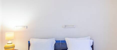 Uma cama de casal com roupa de cama branca, uma luminária de cabeceira e um pequeno objeto decorativo na mesinha de cabeceira.
