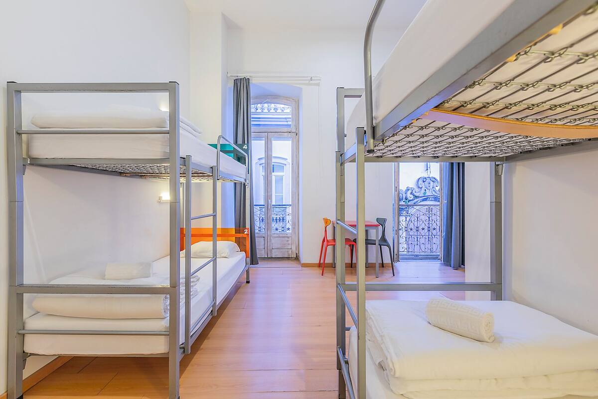 Ein Schlafsaalzimmer mit Stockbetten, einem kleinen Tisch, Stühlen und Blick durch eine Tür.