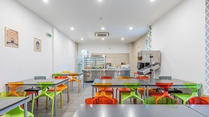 A modern dining area with colorful chairs and tables, a kitchen in the background, and a clock on the wall.