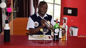 A person preparing a drink at a bar with a cutting board, a knife, and various bottles.