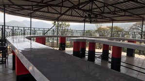 A covered outdoor seating area with a long counter, metal railings, and a view of distant hills.