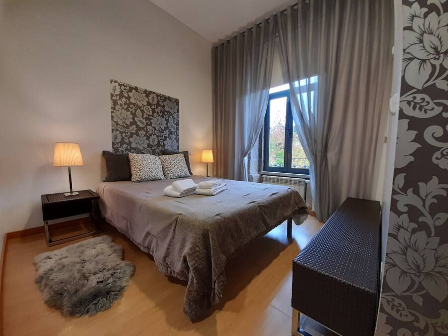 A bedroom with a bed, two bedside lamps, a radiator, a window with curtains, a fluffy rug, and a wall with a floral pattern.