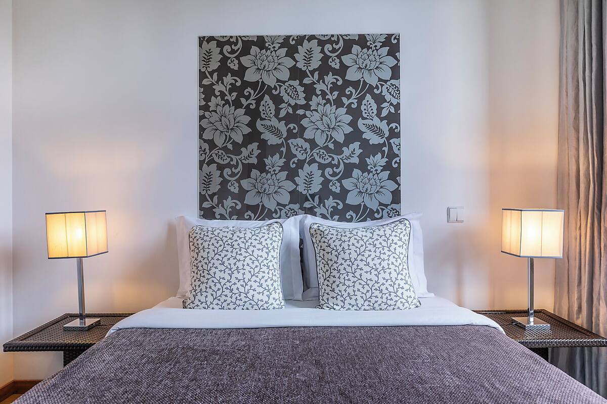 Quarto de hotel com uma cama, duas mesas de cabeceira, uma luminária de parede e um papel de parede estampado.