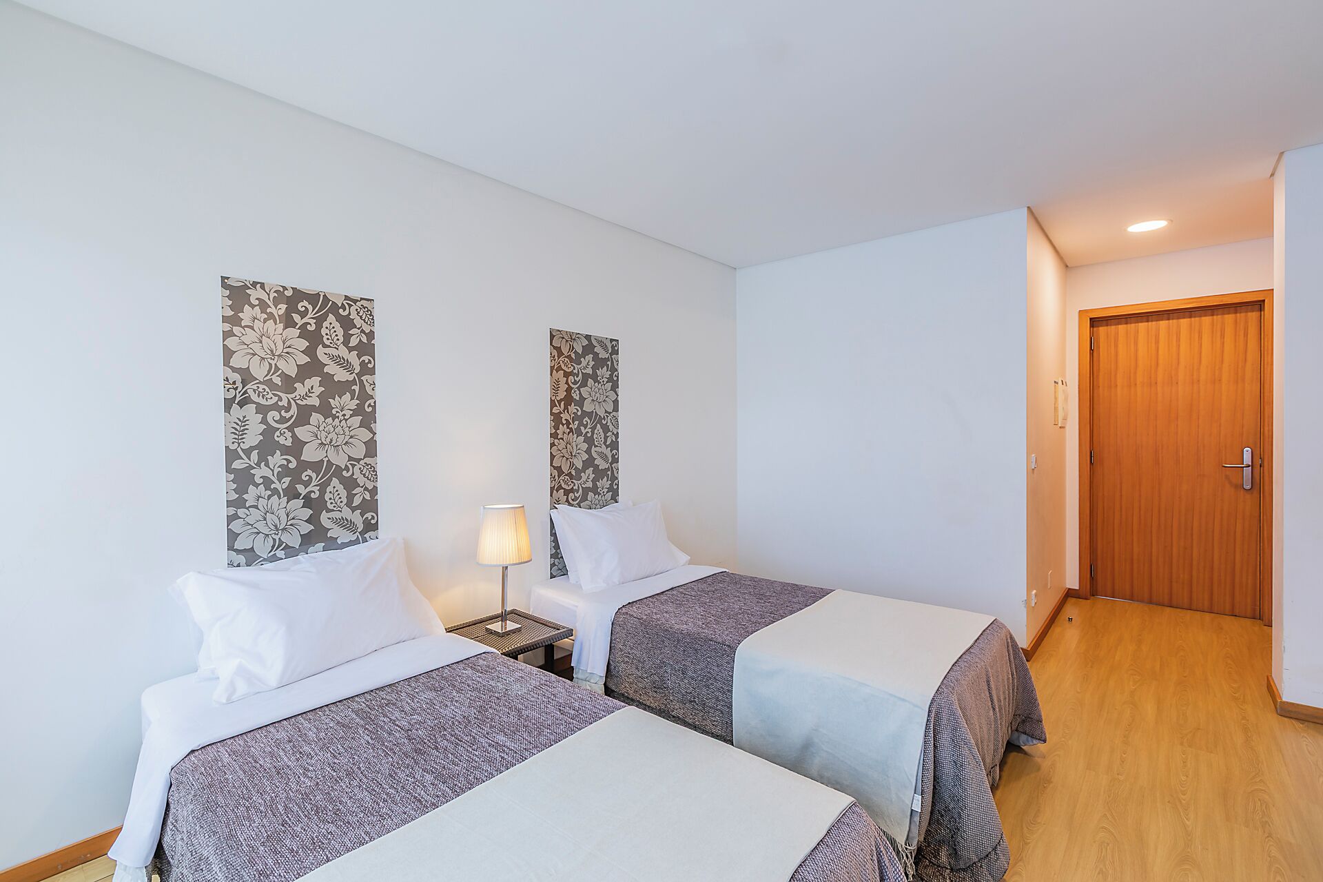 Una habitació d'hotel amb dos llits, una taula de nit amb una làmpada, una porta de fusta i paper pintat amb motius florals.