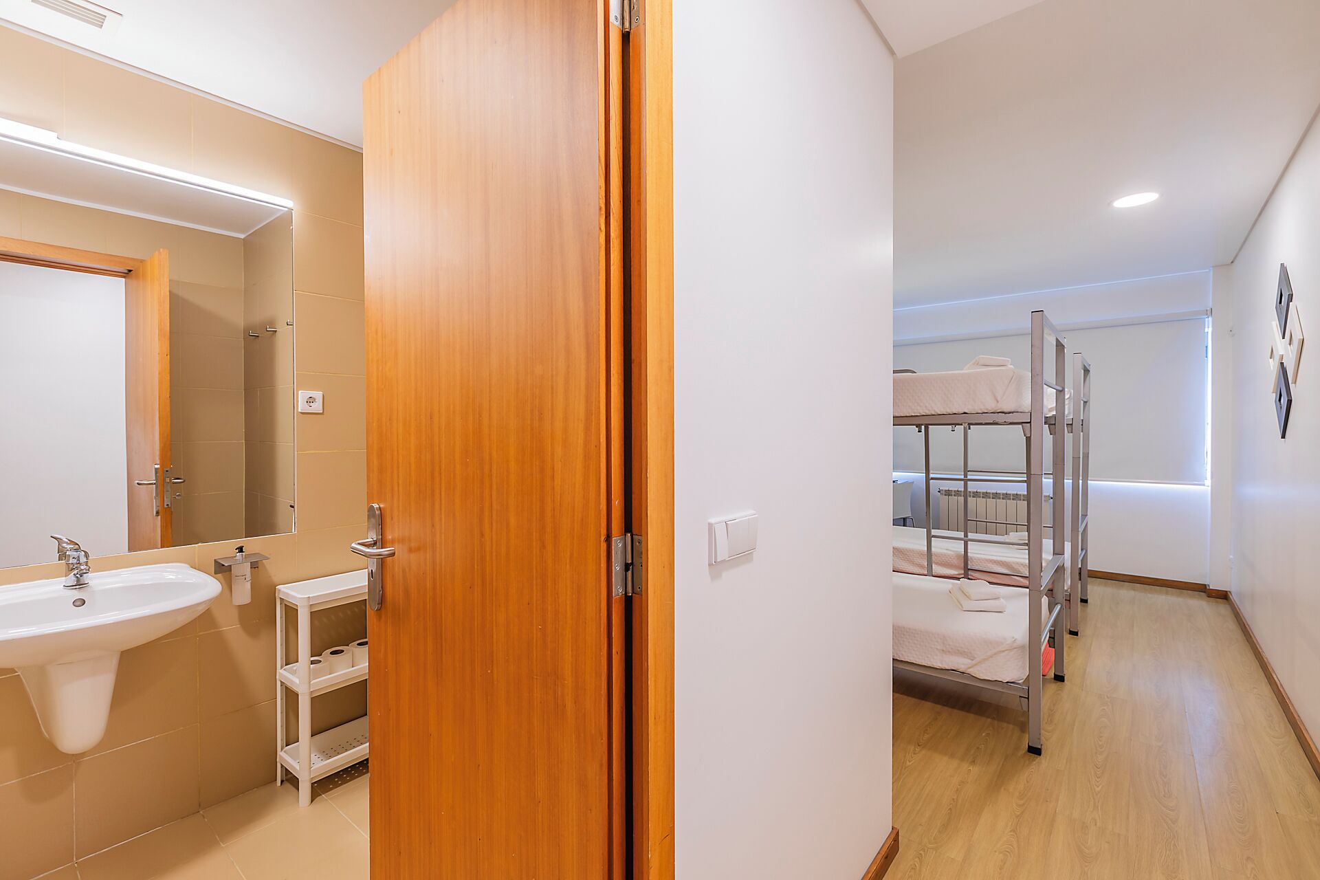 A hotel room with a wooden door, a sink, a mirror, a shelf, a bunk bed, and a radiator.