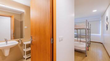 A hotel room with a wooden door, a sink, a mirror, a shelf, a bunk bed, and a radiator.
