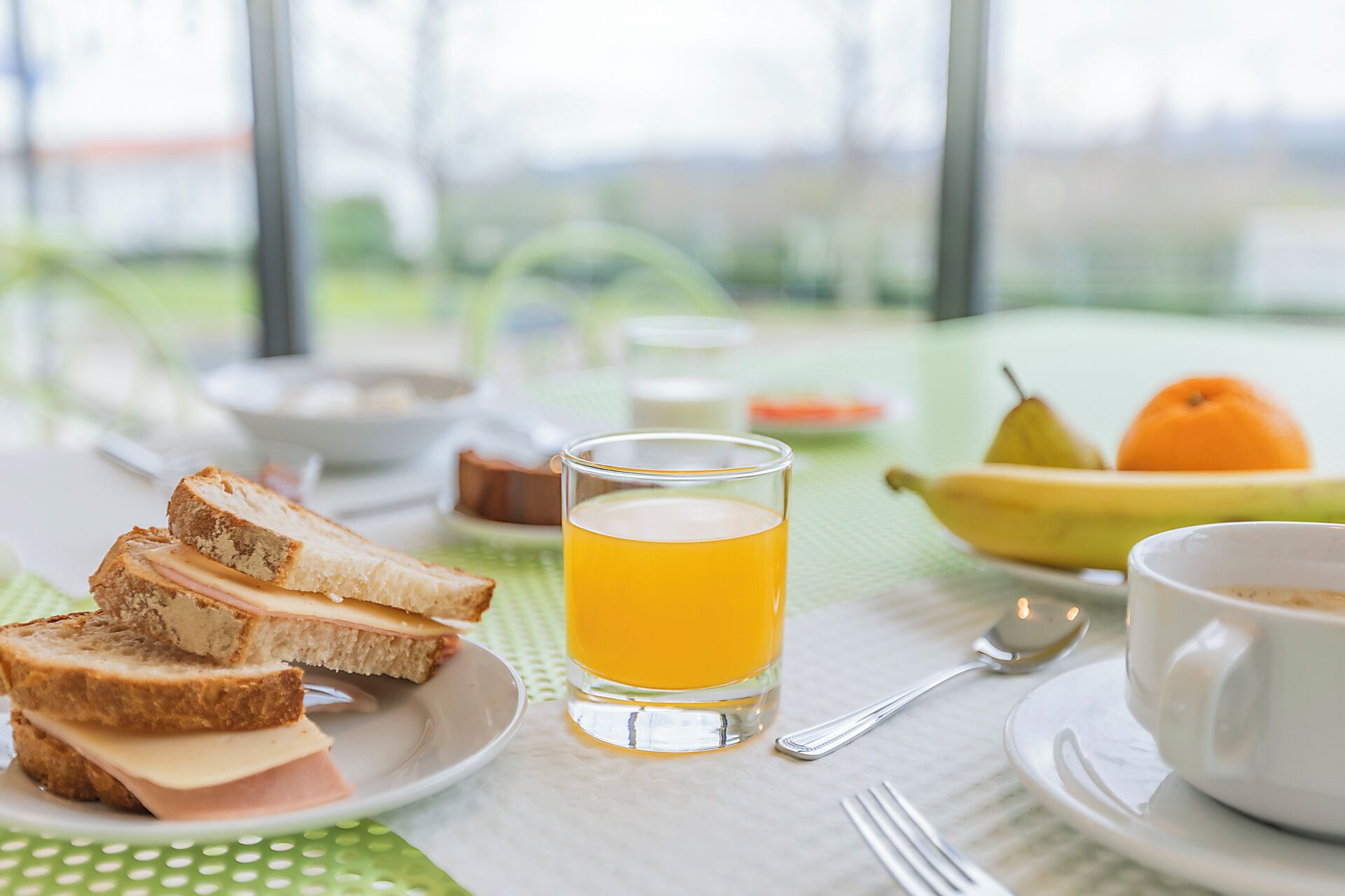 A breakfast setting with toast, a glass of orange juice, a bowl of cereal, a banana, an orange, and a cup of coffee on a table with a view outside.