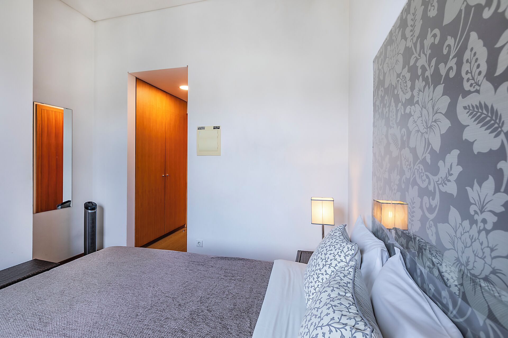A hotel room with a bed, two bedside lamps, a wooden wardrobe, a mirror, and a floral-patterned wall.