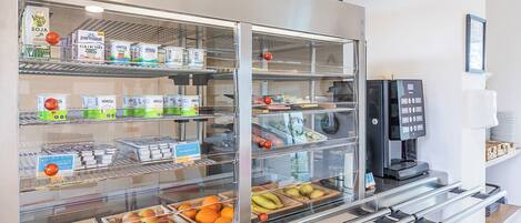 Una estación de desayuno autoservicio con vitrinas refrigeradas, una máquina de café y una variedad de alimentos.
