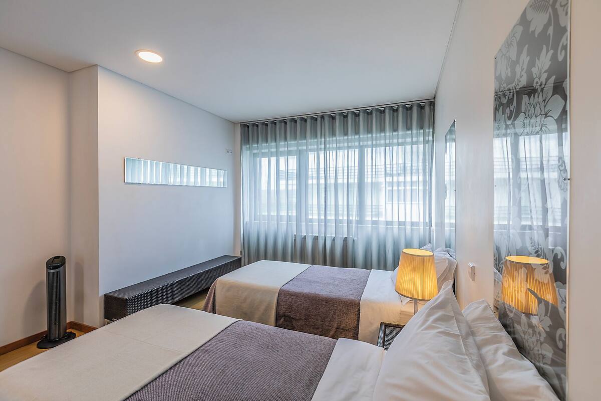 Quarto de hotel com duas camas, um aquecedor portátil, radiador e uma janela grande com cortinas.