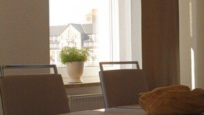 Et spiseområde med et bord, stole, en potteplante og udsigt til bygninger gennem et vindue.