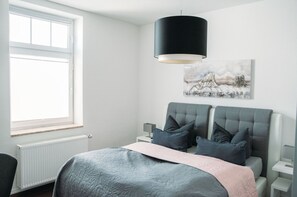 A bedroom with a bed, two bedside tables, a radiator, a window, and a hanging lamp.