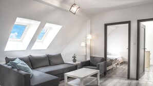 Une chambre de style loft moderne, avec un plafond en pente, des fenêtres de toit, un canapé gris, une table basse blanche et un lit doté d’une tête de lit en bois.