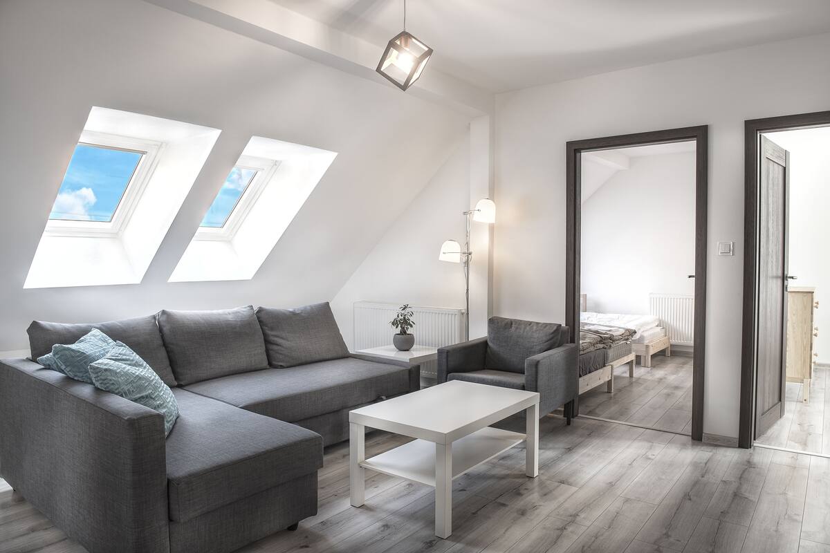 A modern loft bedroom with a sloped ceiling, skylights, a grey sofa, a white coffee table, and a bed with a wooden headboard.