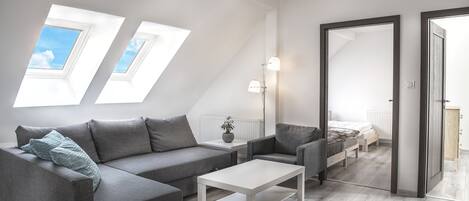 Un dormitorio tipo loft moderno con techo inclinado, tragaluces, un sofá gris, una mesa de centro blanca y una cama con cabecero de madera.