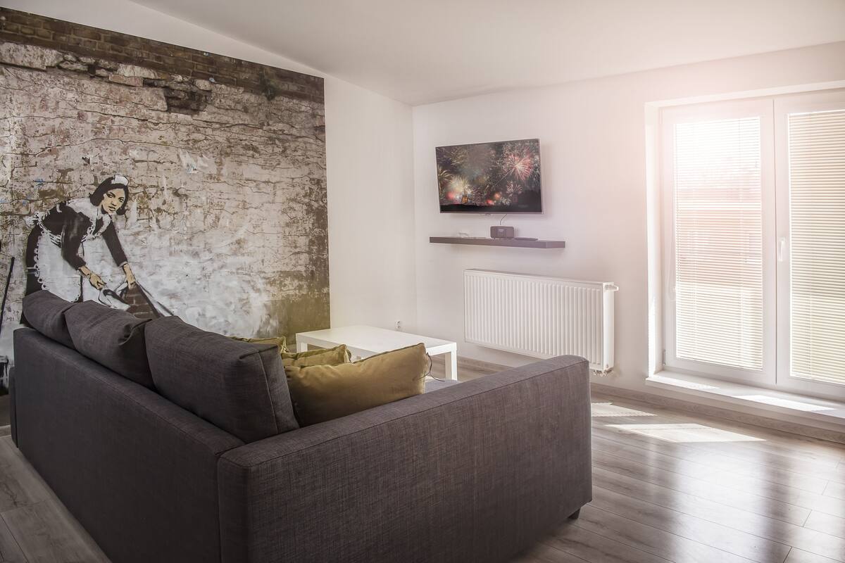 A modern living room with a grey sofa, a white coffee table, a mural on the brick wall, a flat-screen TV on a shelf, and a large window with blinds.