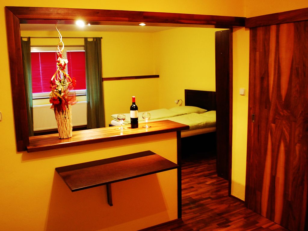 Una habitación pequeña con un banco de madera, una cama, una mesita de noche con una botella y vasos, y un espejo que refleja una ventana con cortinas.
