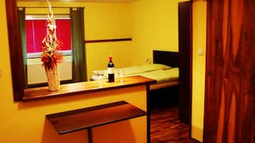 Una habitación pequeña con un banco de madera, una cama, una mesita de noche con una botella y vasos, y un espejo que refleja una ventana con cortinas.