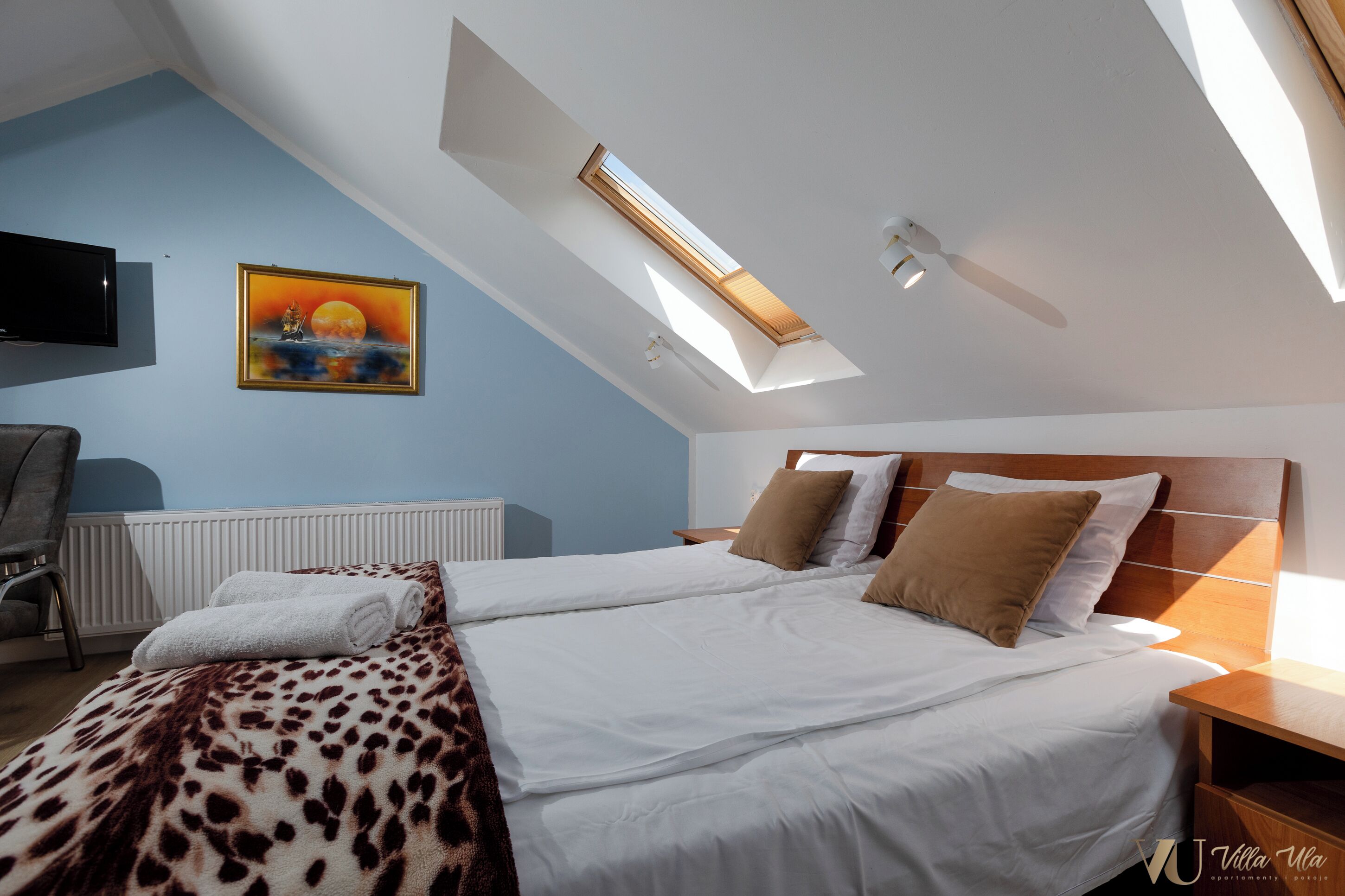 Ein Schlafzimmer mit Dachfenster, einem Bett mit weißer Bettwäsche, einer Decke mit Leopardenmuster, einem Gemälde von einem Sonnenuntergang und einem an der Wand befestigten Fernseher.