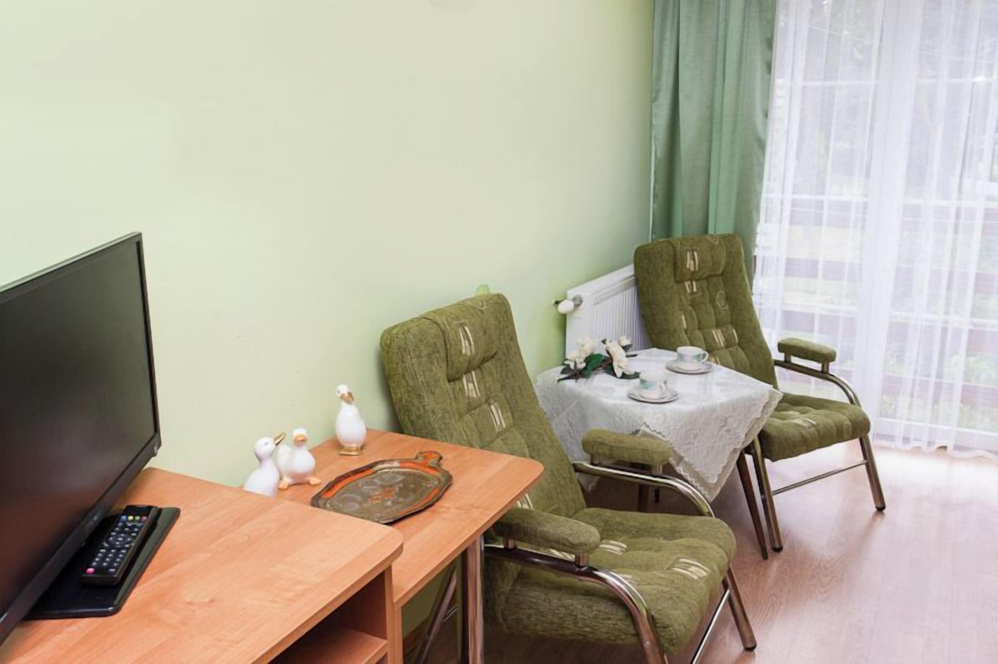 A room with a TV, a wooden table, two green upholstered chairs, a small table with a white tablecloth, and a window with sheer curtains.