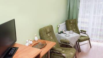 A room with a TV, a wooden table, two green upholstered chairs, a small table with a white tablecloth, and a window with sheer curtains.