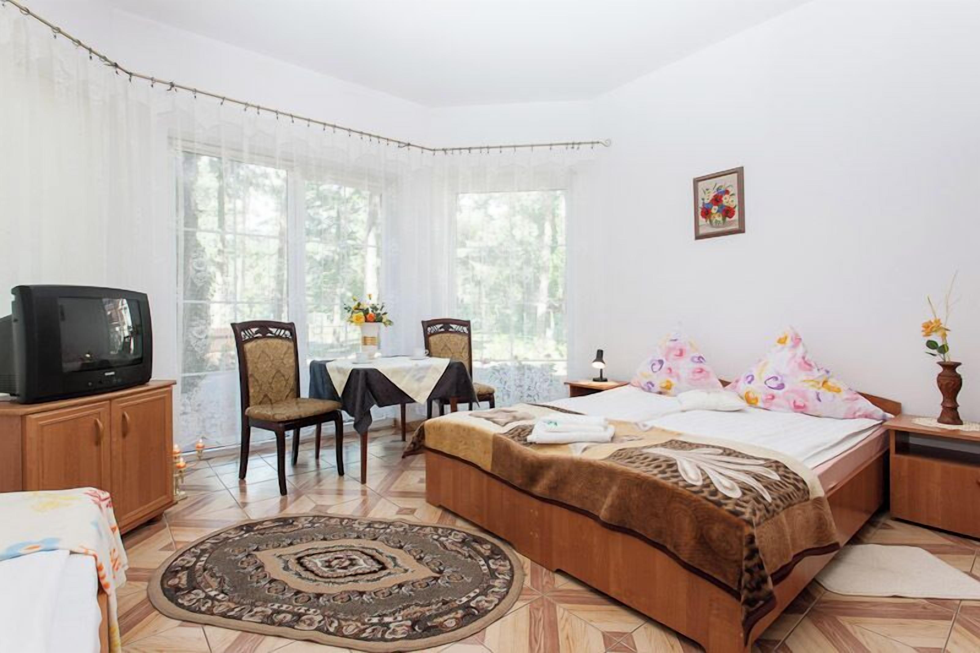 A bedroom with a bed, a television, a dining table with chairs, and a window with curtains.