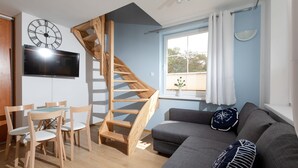 Un salon moderne avec un escalier en bois, un canapé gris, des chaises en bois, un téléviseur à écran plat et une grande horloge murale.