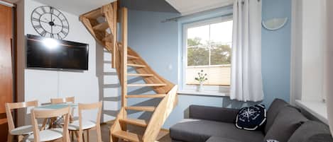 A modern living room with a wooden staircase, a grey sofa, wooden chairs, a flat-screen TV, and a large wall clock.