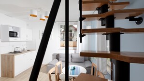 Une cuisine moderne avec des armoires blanches, un four à micro-ondes et une cuisinière à gaz. On y trouve un coin repas avec une table ronde et des chaises, ainsi qu’un canapé gris. Un escalier avec des marches en bois et des rampes noires est visible.