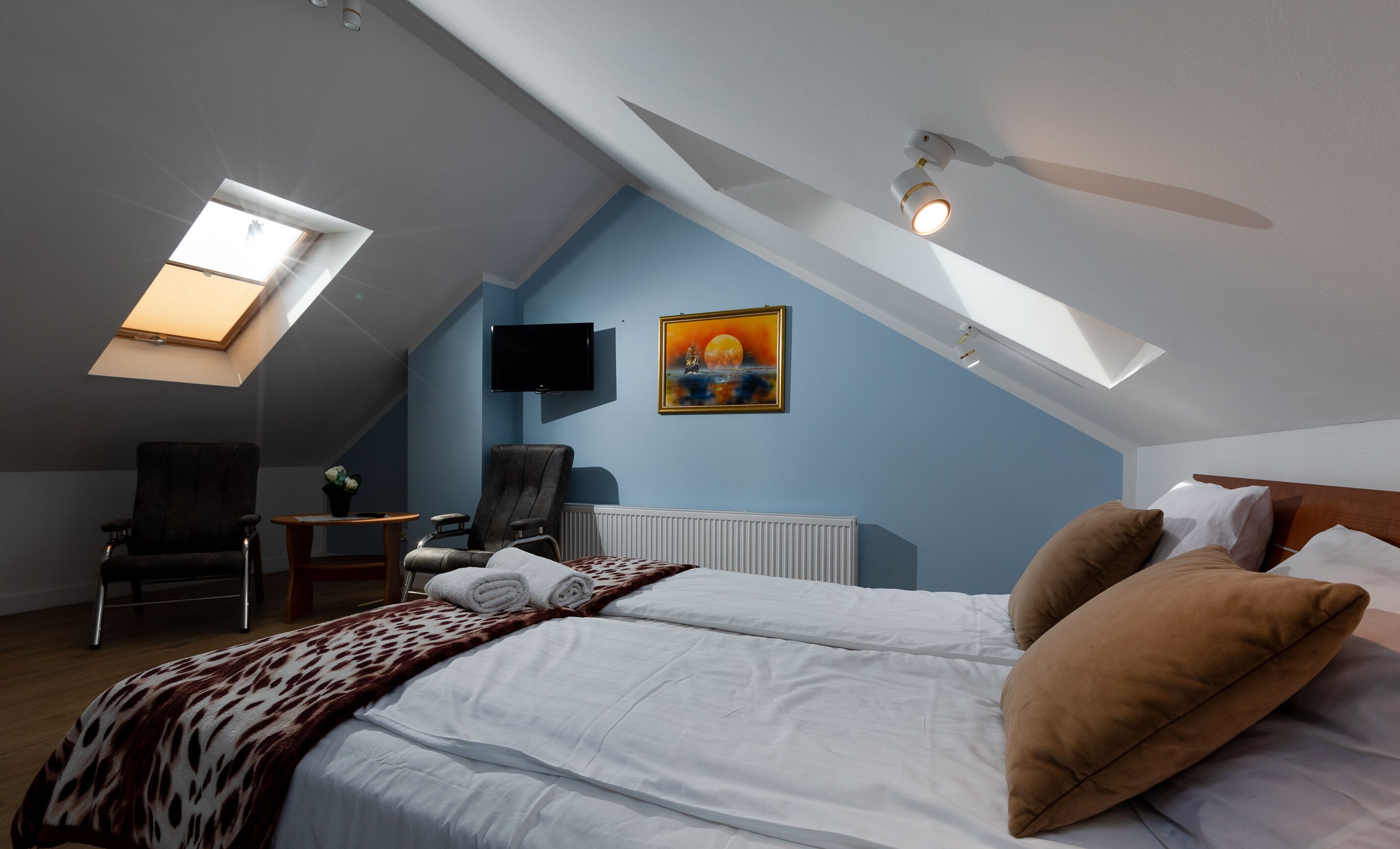 A bedroom with a sloped ceiling, a bed with white linens, a leopard print blanket, a brown pillow, a chair, a small table, a painting, and a mounted TV.