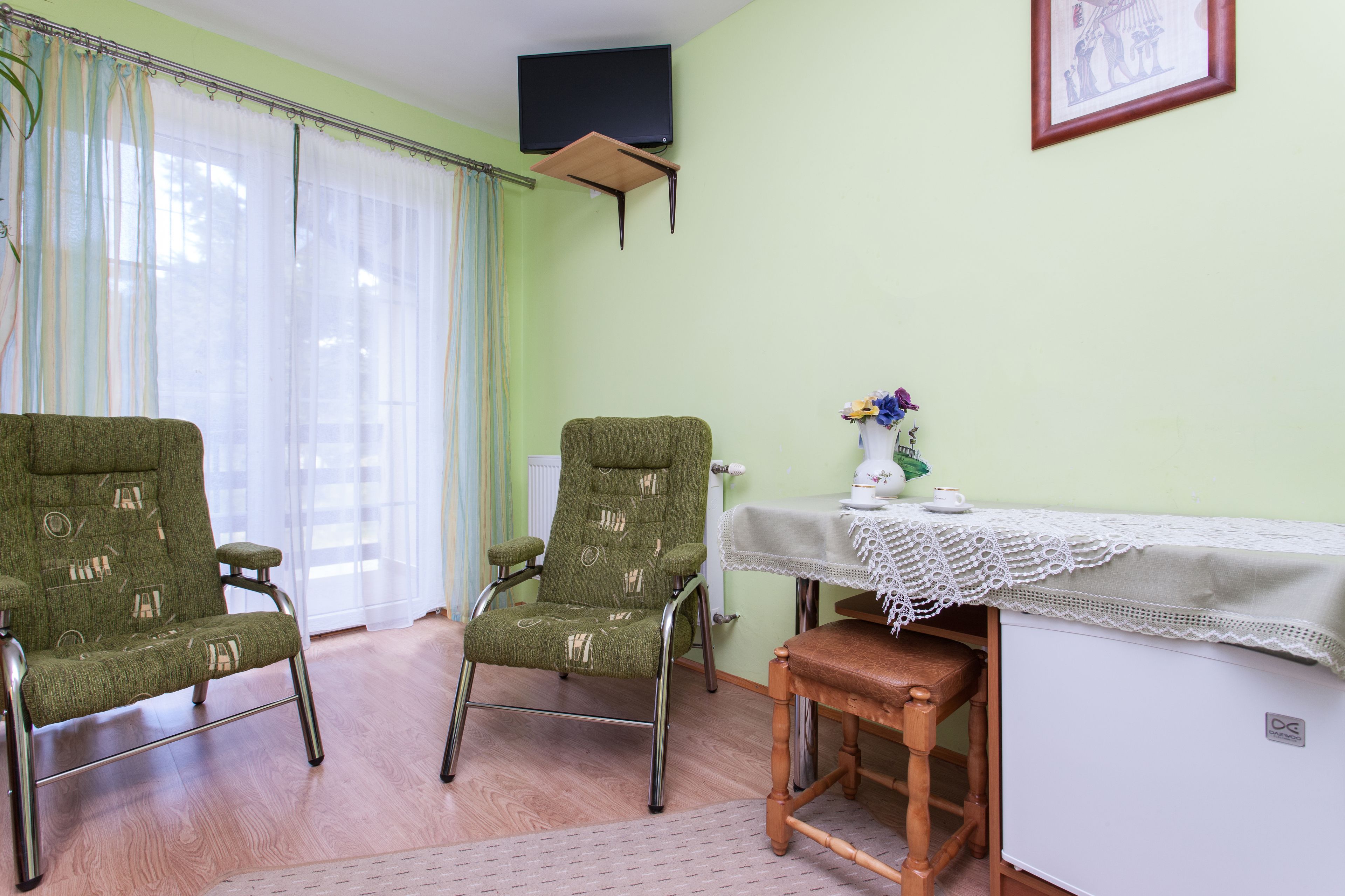 A room with two green armchairs, a wooden table with a lace tablecloth, and a white cabinet.
