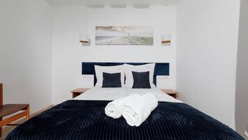 Un letto rifatto con cura, con una testiera blu scuro, lenzuola bianche e un asciugamano bianco arrotolato. Sopra il letto, un quadro raffigura una spiaggia.