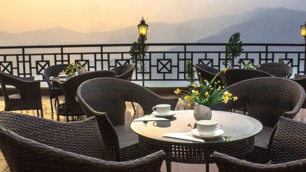 An outdoor seating area with wicker furniture and a table set for two, overlooking a mountainous landscape. - Daizzo Maple Residency (Gangtok)