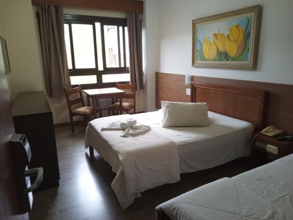 A hotel room with a bed, a desk, and a chair. There is a painting on the wall and a window with curtains.