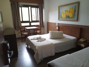 A hotel room with a bed, a desk, and a chair. There is a painting on the wall and a window with curtains.