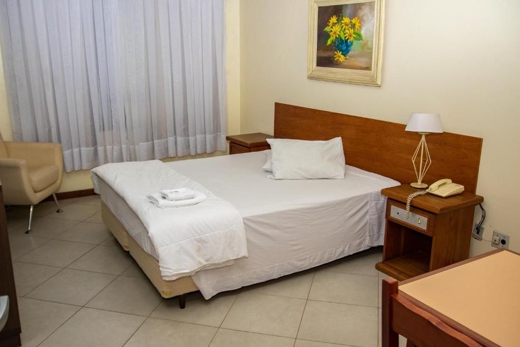Una habitación de hotel con cama, una silla, una mesita de noche con lámpara y un cuadro en la pared.
