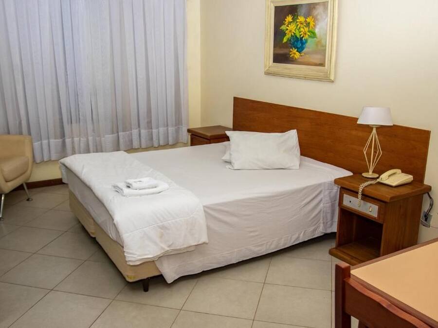 Una habitación de hotel con cama, una silla, una mesita de noche con lámpara y un cuadro en la pared.