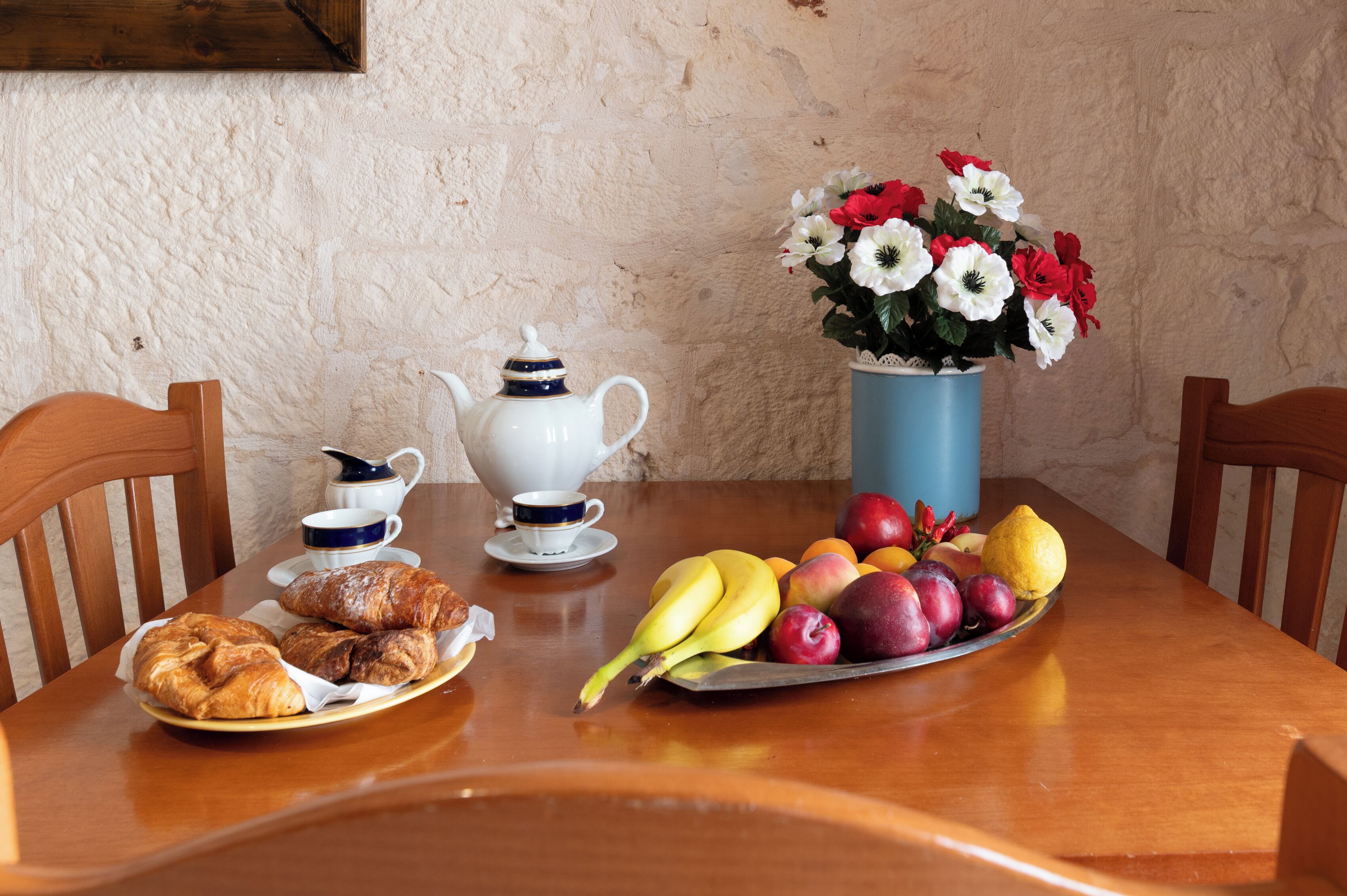 Una mesa de madera con un plato de cruasanes, un frutero, una tetera, tazas y un jarrón de flores.