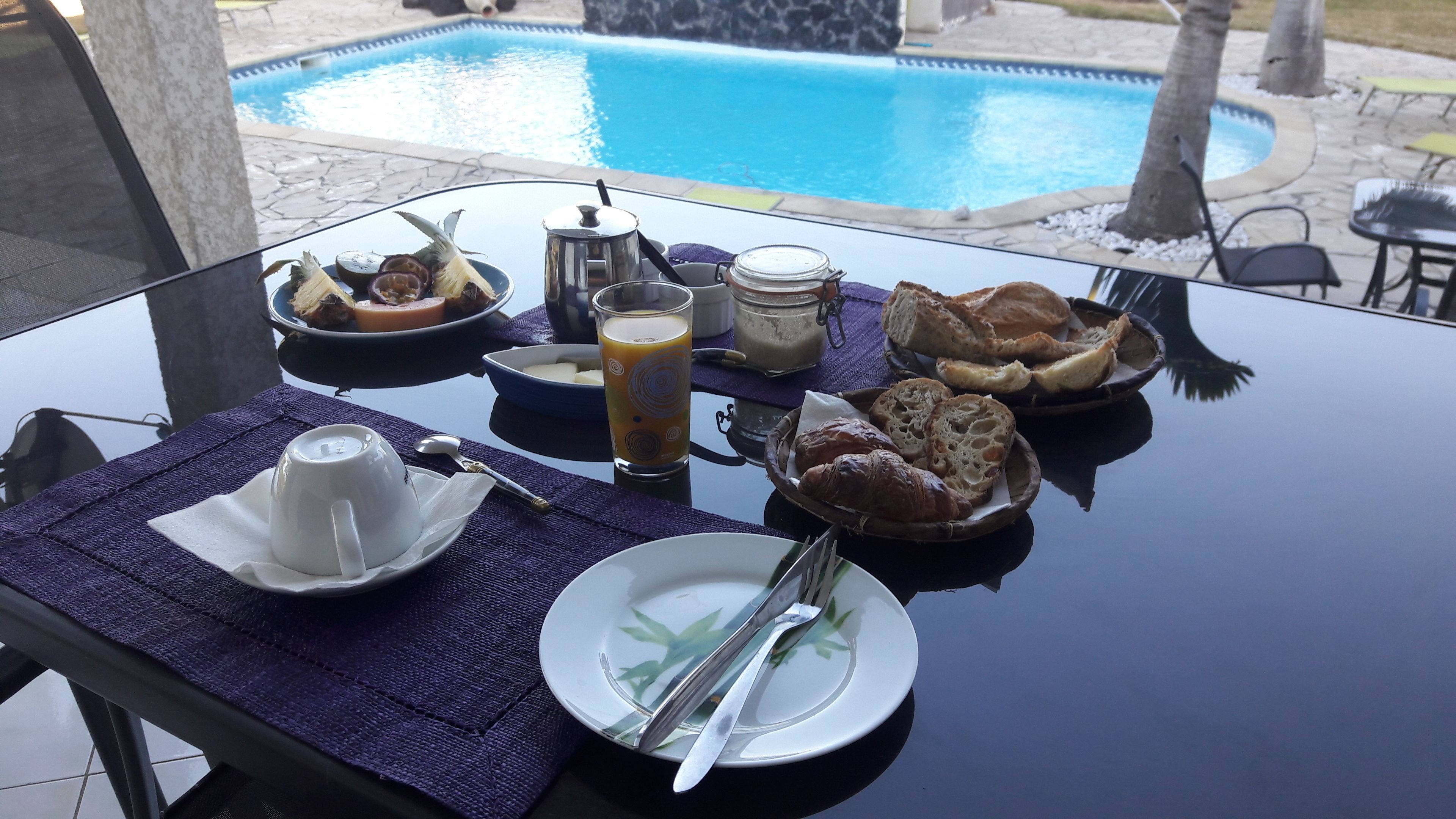 A breakfast table with a variety of food items, a glass of orange juice, and a view of a swimming pool in the background.