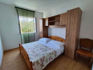 A single bed with a floral quilt, a wooden headboard, a small wooden cabinet above it, and a chair to the right.