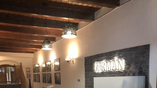 A reception area with a wooden reception desk, a stone wall with the name 'GURMAN' illuminated, and a staircase in the background. - Hotel Gurmán (Horšovský Týn)